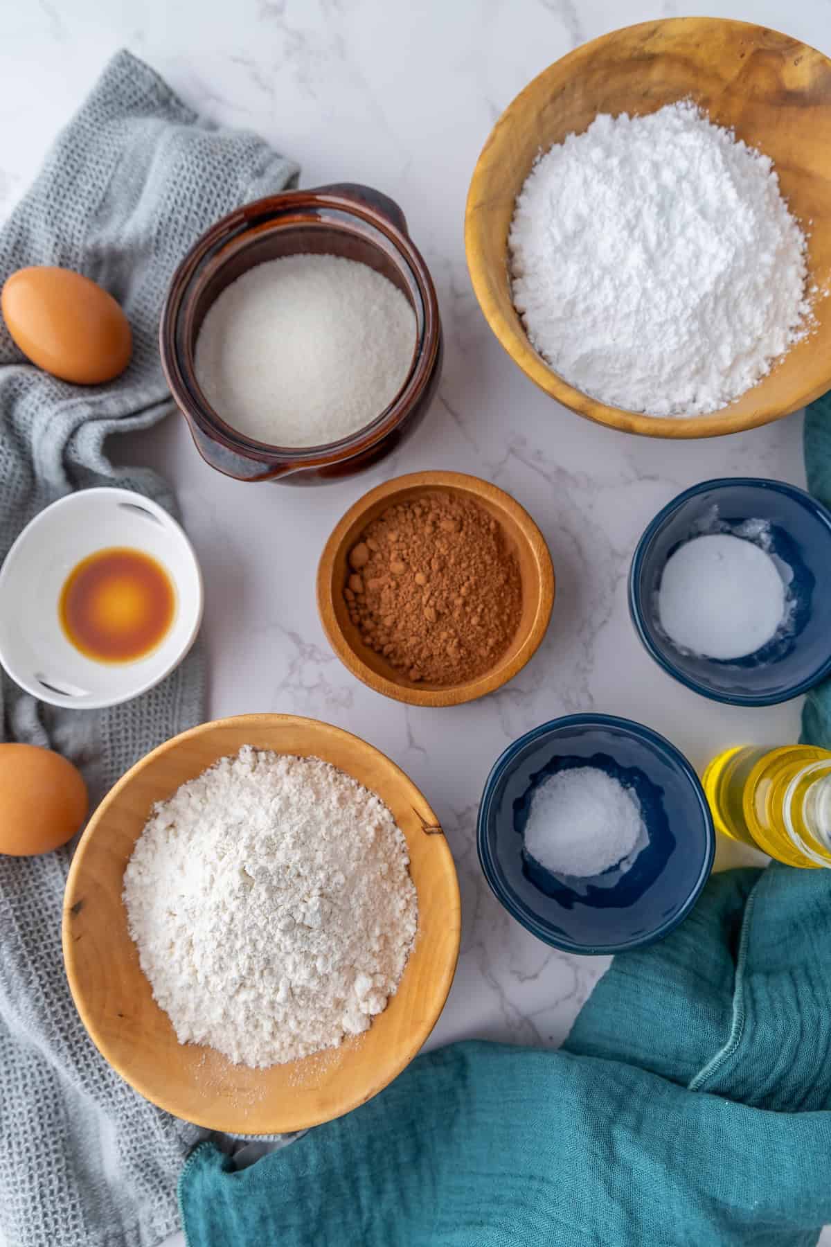 Baking ingredients for chocolate crinkle cookies are artfully arranged on a marble surface: flour, sugar, cocoa powder, eggs, vanilla, oil, and a pinch of salt in various bowls and containers.