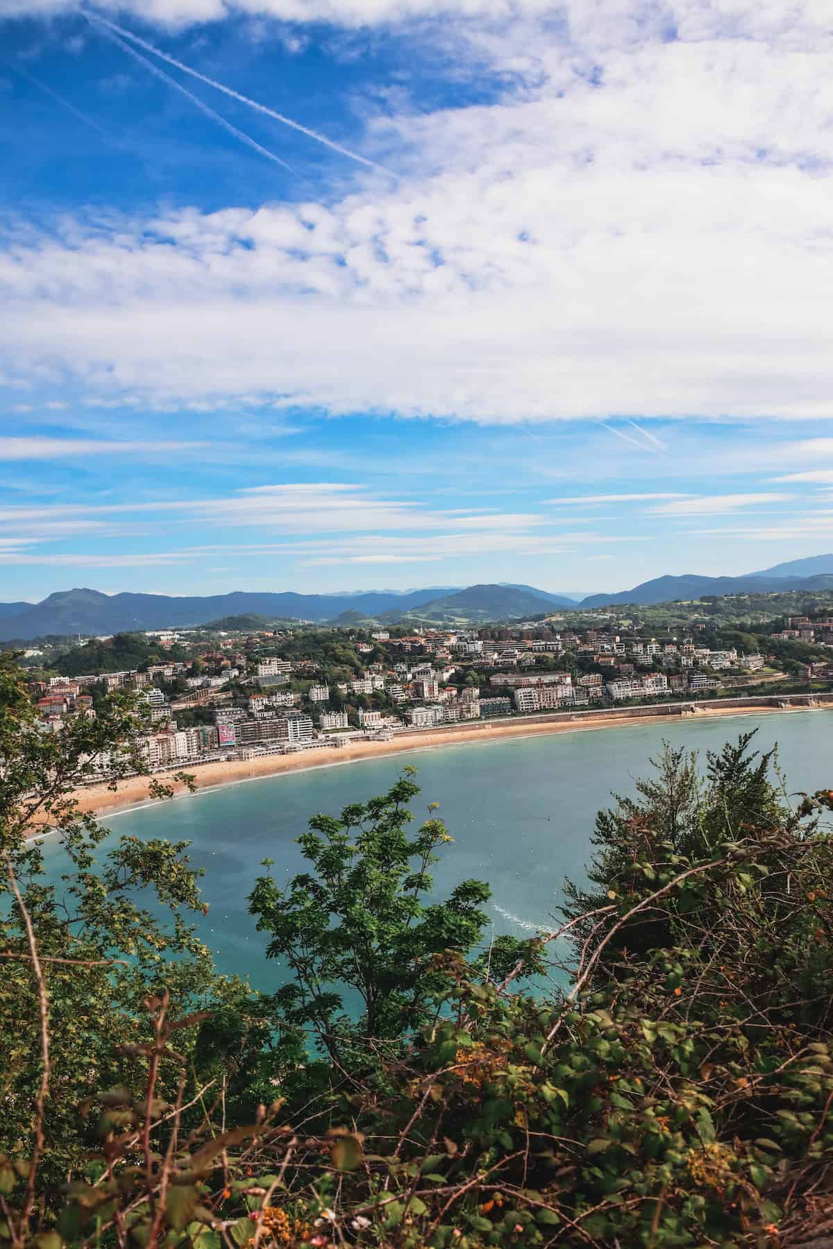 A coastal city with mountains in the distance is seen from a high vantage point. Trees and vegetation are in the foreground, and the sky is partly cloudy with some aircraft contrails visible.